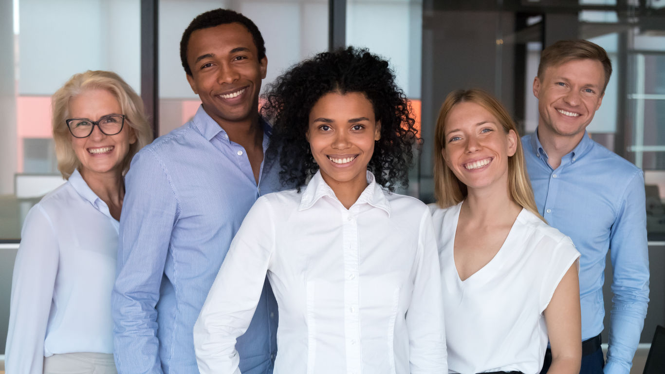 Attractive successful people standing together smiling posing looking at camera feels proud and satisfied. Multi-ethnic businesspeople led by afro mixed race woman photographing in modern office room